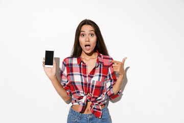Fototapeta premium Amazed young pretty girl in checkered shirt showing thumb up gesture and holding smartphone