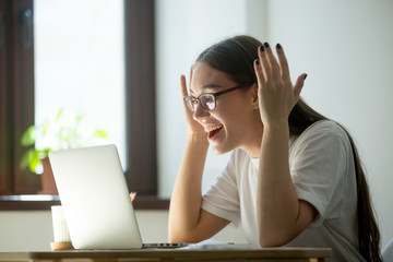 Young adult woman receiving good news via her laptop computer, reading email, videochatting with...