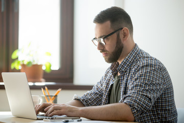 Millennial generation man in glasses and casual clothing working on laptop computer to solve a problem, concentrating on finding best solution.