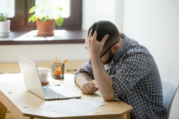 Depressed man looking in his laptop computer receiving bad news. His hand holding his head in sad gesture. Bankruptcy or dismissal concept.  Side view.