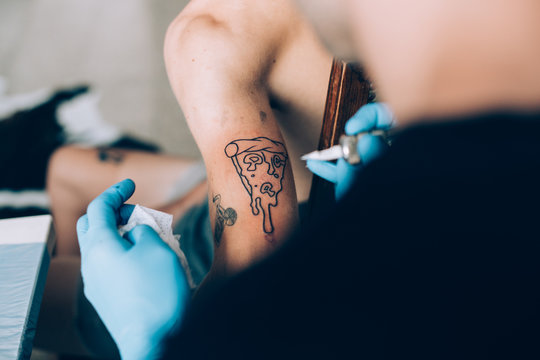Close Up Soft Focus Shot Of Professional Tattoo Artist Tattooing Funny And Hillarious Pizza Art On Arm Of Young Hipster Man, Wears Hygienic Blue Latex Gloves