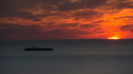 Naklejka na ściany i meble A container ship passes beneath the setting sun near Hout Bay, South Africa