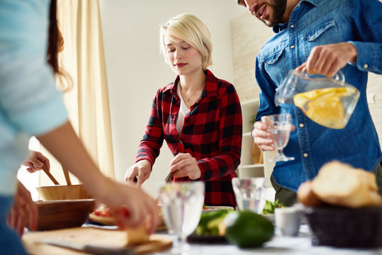 Group of modern young people standing at big table with food on it preparing dinner together for friends gathering at home, cutting vegetables and poring drinks