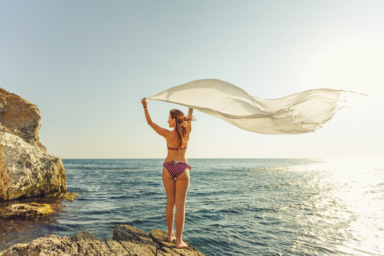 Silhouette of a beautiful woman relaxing on the beach. The girl gladly raises her hands and the sarong pareo develops in the wind against the blue sky. The concept of summer vacations and travel.