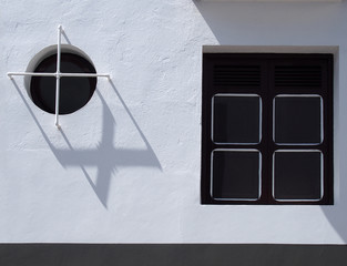 two windows in modern house square and round with stell bars and closed shutters on a white wall with sunlight and shadows
