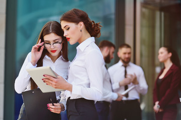 two young pretty girls or business women in office style clothes with a tablet in their hands discuss the plan against the backdrop of colleagues or employees of the company