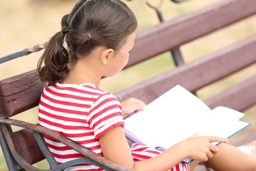 Cute little girl reading book while sitting on bench in park