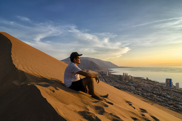 Young man sitting at the summit of enormours sand dune and overlooking city and ocean at the sunset