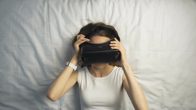 Dark hair caucasian girl puts on her virtual reality glasses, lied down on the huge white bed, slow motion
