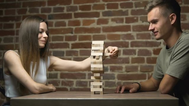 Two caucasian keen friends playing jenga at the table next to the brick wall, the tower falls, slow motion