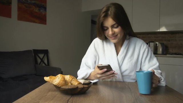 Pretty caucasian dark hair girl, in a white bathrobe, having her morning coffee while using smartphone in the kitchen, slow motion
