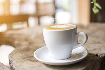 White cup with steaming hot cappuccino on the wooden table table with blurred background at coffee shop