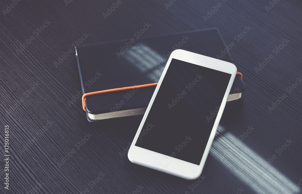 Poster Blank screen of mobile phone lay on black notebook near window on black wooden modern desk at afternoon sunlight, Mock up for display of design or content,Clipping path smart phone screen.