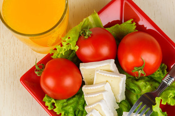 Tomatoes and cheese slices lie on a sheet of fresh salad in a red plate. A glass of orange juice stands side by side on the table.