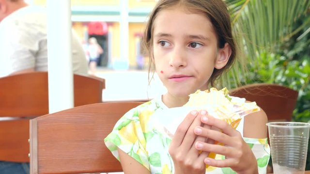 An attractive girl eats fresh pita bread with a salad of fresh vegetables and meat, sitting in a fast food cafe under a tree in the park. 4K slow motion