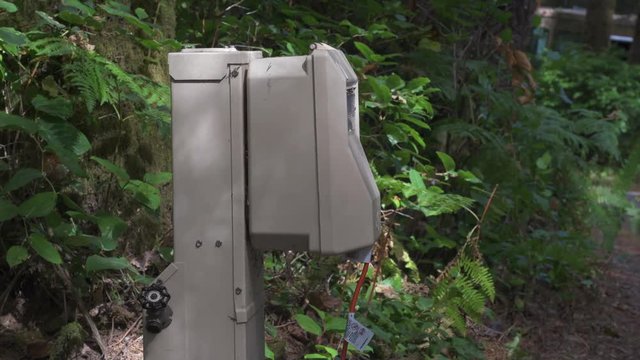 At a green, forested campsite, a person lifts the cover of a power hookup box, unplugging a orange extension cord, and closes the lid quickly.