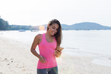 Fototapeta premium Portrait Of Young Woman Runner On Beach Holding Cell Smart Phone While Jogging On Seaside Female Happy Smiling Girl Sport Runner Healthy Lifestyle Concept