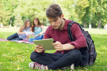 Student life. Young male student reads your electronic digital tablet sitting on the green lawn near the university.