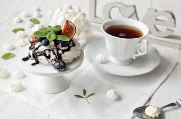 A cup of tea and a meringue cake decorated with blueberries and figs on a white wooden background. Selective focus, close-up.