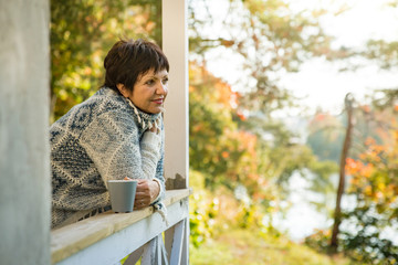 Mature attractive woman standing on terrace with cup of hot coffee wrapped up in knitted warm sweater. Lake house in autumn, yellow and red leaves on trees. Cozy sunny day