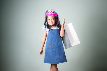 pretty  little asian girl in blue jeans dungarees holding shopping bags with white concrete wall background.portrait of young kid smiling and looking at camera