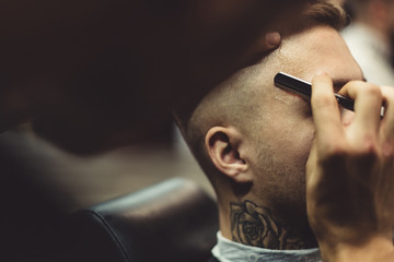 Anonymous barber shaving stylish tattooed man with razor in barbershop.