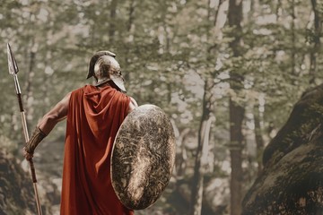 Back view of man in gladiator helmet and red long cloak standing with round shield and spear in hands looking away in woods. Spartan. - obrazy, fototapety, plakaty