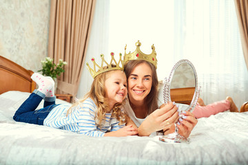 Mother and daughter in crowns play with a mirror on the roof near the window in the room. Mothers Day. - obrazy, fototapety, plakaty