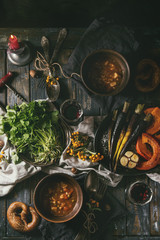 Fall holiday table decoration setting with bowls of hot carrot potato soup, baking pumpkin, carrot, garlic, fresh coriander, pretzels bread, red wine, berries and candle. Flat lay over wooden table