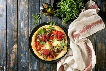 Crédence de cuisine en verre imprimé Pizzeria Whole homemade pizza with cheese and bresaola, served on black plate with fresh arugula, olive oil and kitchen towel over old wooden plank background. Flat lay.