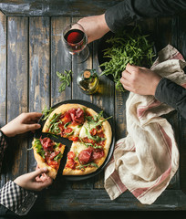 Handen nemen gesneden zelfgemaakte pizza met kaas en bresaola, geserveerd op zwarte plaat met verse rucola, olijfolie, glas rode wijn en keukenhanddoek over oude houten plank achtergrond. Plat leggen.