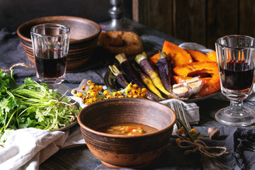 Fall holiday table decoration setting with bowls of hot carrot potato soup, baking pumpkin, carrot, garlic, fresh coriander, pretzels bread, red wine, berries, candle over wooden table. Rustic style