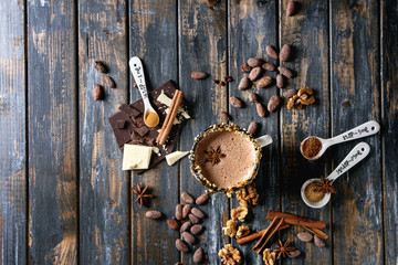 Vintage mug of hot chocolate, decor with nuts, caramel, spices. Ingredients above. Chopped dark and white chocolate, cocoa beans, anise over old wooden table. Top view with space. Dark rustic style