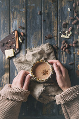 Female hands hold vintage mug of hot chocolate, decorated with nuts, caramel, spices. Ingredients above over old wooden table. Flat lay with space. Dark rustic style