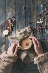 Photo sur Plexiglas Chocolat Female hands hold vintage mug of hot chocolate, decorated with nuts, caramel, spices. Ingredients above over old wooden table. Flat lay with space. Dark rustic style
