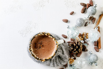 Vintage mug in wool scarf of hot chocolate, decor with nuts, caramel, spices. Ingredients and Christmas toys above over white texture background. Top view with space.