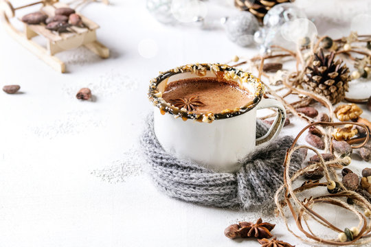 Vintage mug in wool scarf of hot chocolate, decor with nuts, caramel, spices. Ingredients and Christmas toys above over white texture background with space.