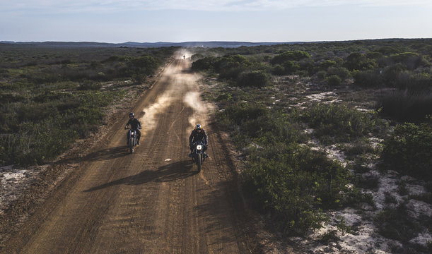 Landscape with two men riding cafe racer motorcycles in circles on a dusty dirt road.
