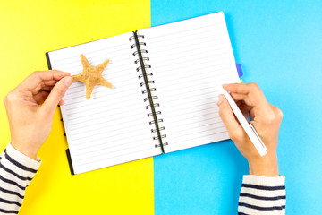 Vacation planning concept. Woman hand writing in open travel notebook over blue and yellow background.