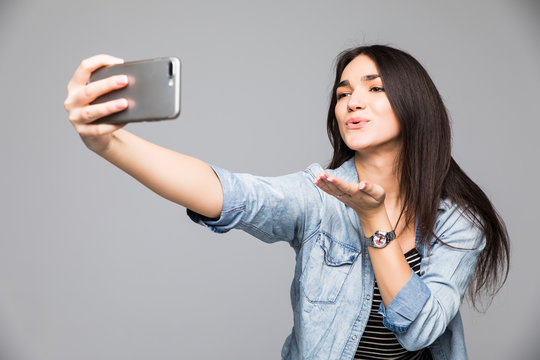 Beautiful brunette woman making a selfie blowing a kiss holding the smartphone isolated on a gray background