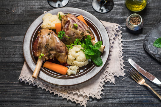 Lamb shank dinner on slate tray with vegetables, parsley and mint jam on black rustic wooden table, top view, copy space