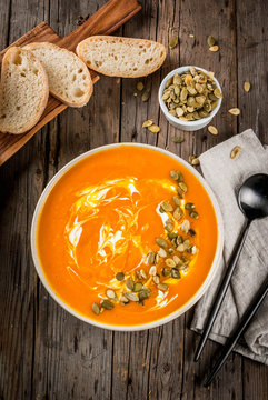 Traditional fall and winter dishes, hot and spicy pumpkin  soup with pumpkin seeds, cream and freshly baked baguette, on old rustic wooden table, copy space top view