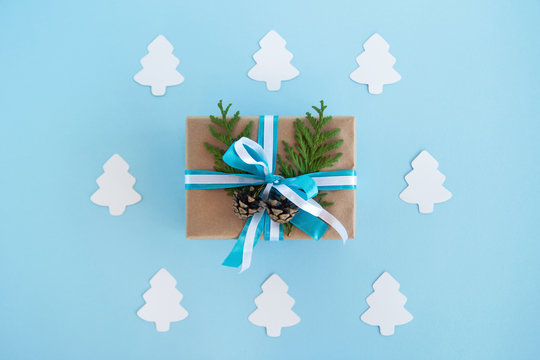 Gift box wrapped of craft paper, blue and white ribbons and decorated fir branches and pinecones on the blue background with white paper fir tree, top view. Christmas present.