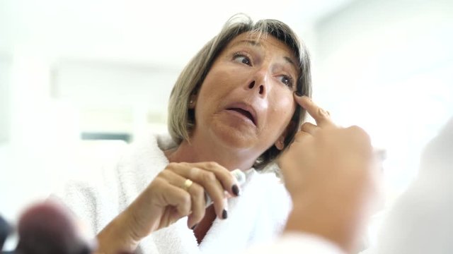 Senior woman in front of mirror applying eye concealer 