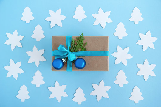 Gift box wrapped of craft paper, blue ribbon and decorated fir branches and blue Christmas balls on the blue background with paper fir tree and star, top view. Christmas present.