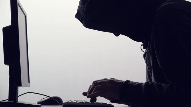 Hooded computer hacker hacking network. Male with black hoodie using computer for identity theft or other criminal online activity.