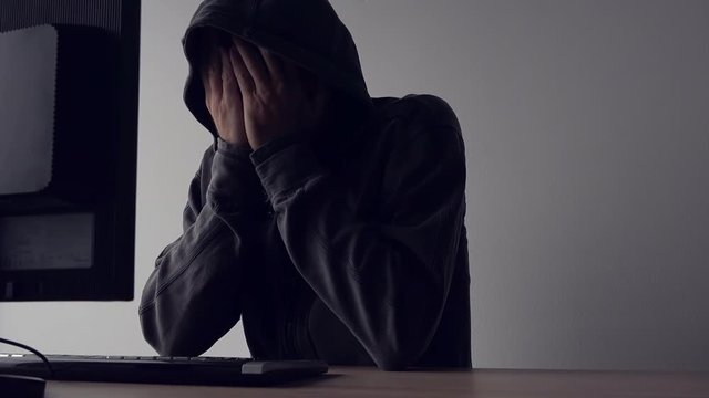 Depressed hooded computer hacker covering face with hands. Male with black hoodie worried for failing in criminal online activity.