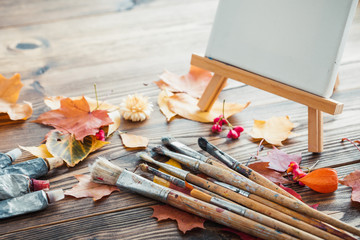 Canvas on easel, paint tubes, brushes for painting and autumn leaves on old wooden background. Retro toned.