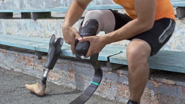 Tilt up of amputee athlete sitting on stadium bleachers and putting on running blade before training