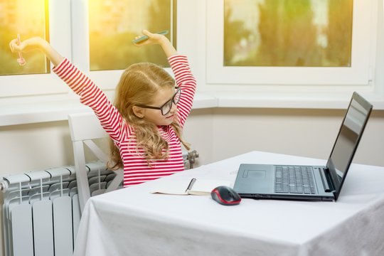 Portrait Of A Cute 7 Year Old Girl Wearing Glasses, Using A Laptop, Smartphone And Writing In A Notepad Sitting At The Table.